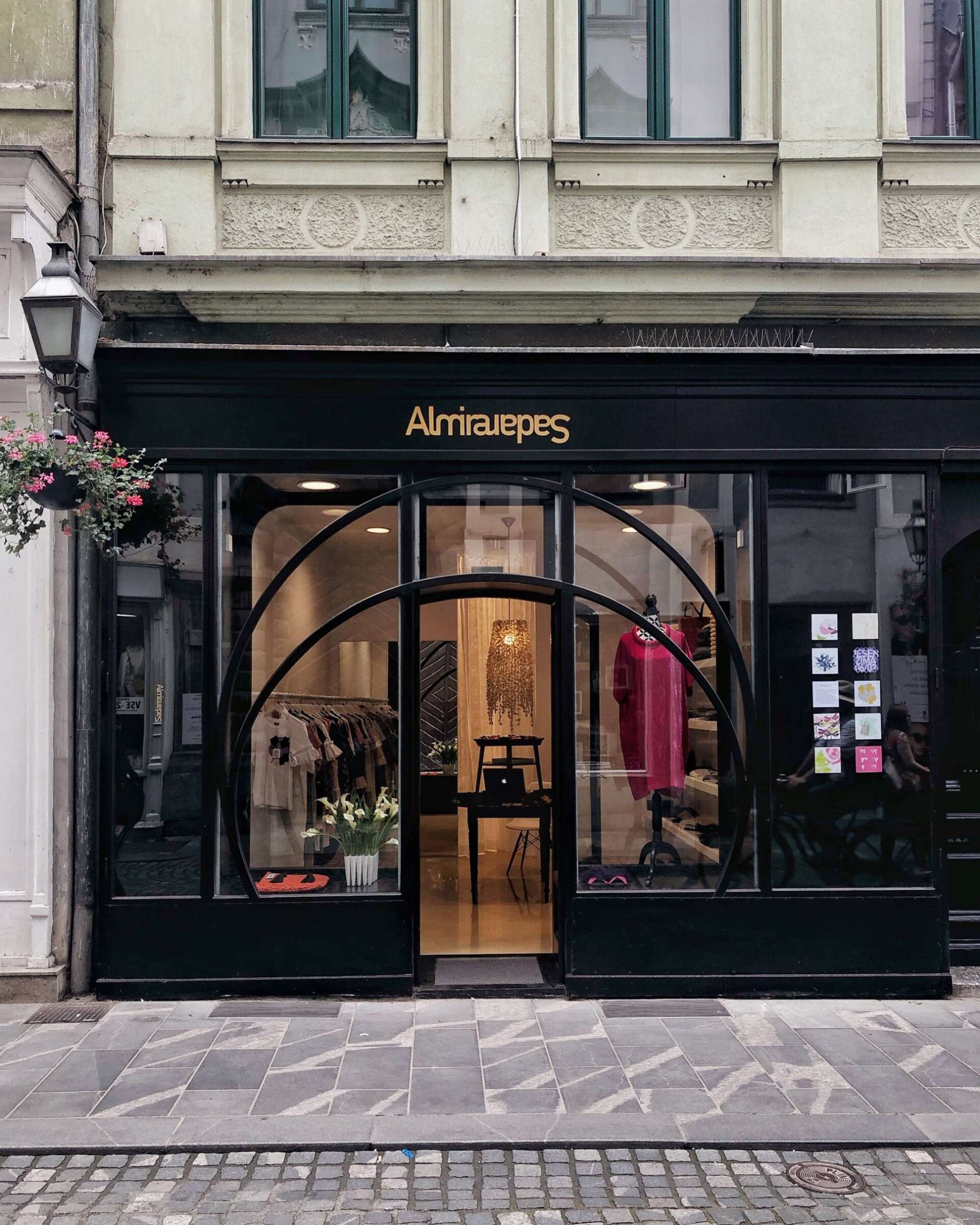 A stylish boutique with large glass windows and elegant curved arches. Inside, a decorative chandelier and a collection of clothing emphasise the elegant setting, and in front of the boutique is a cobbled street.