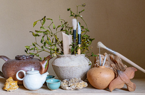 The scene shows a composition of handmade ceramics and a vase filled with sculpting tools and green leaves.