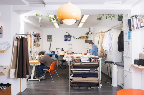 In a bright sewing studio, there are two people at separate workstations, one is sewing with a machine, the other is unravelling fabrics.