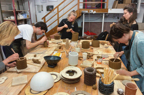 Several people craft pottery in a workshop, shaping and detailing clay. A wooden table holds tools and clay pieces, while shelves in the background store materials.