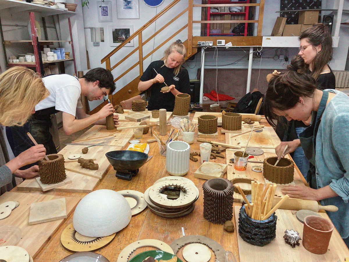 Several people craft pottery in a workshop, shaping and detailing clay. A wooden table holds tools and clay pieces, while shelves in the background store materials.