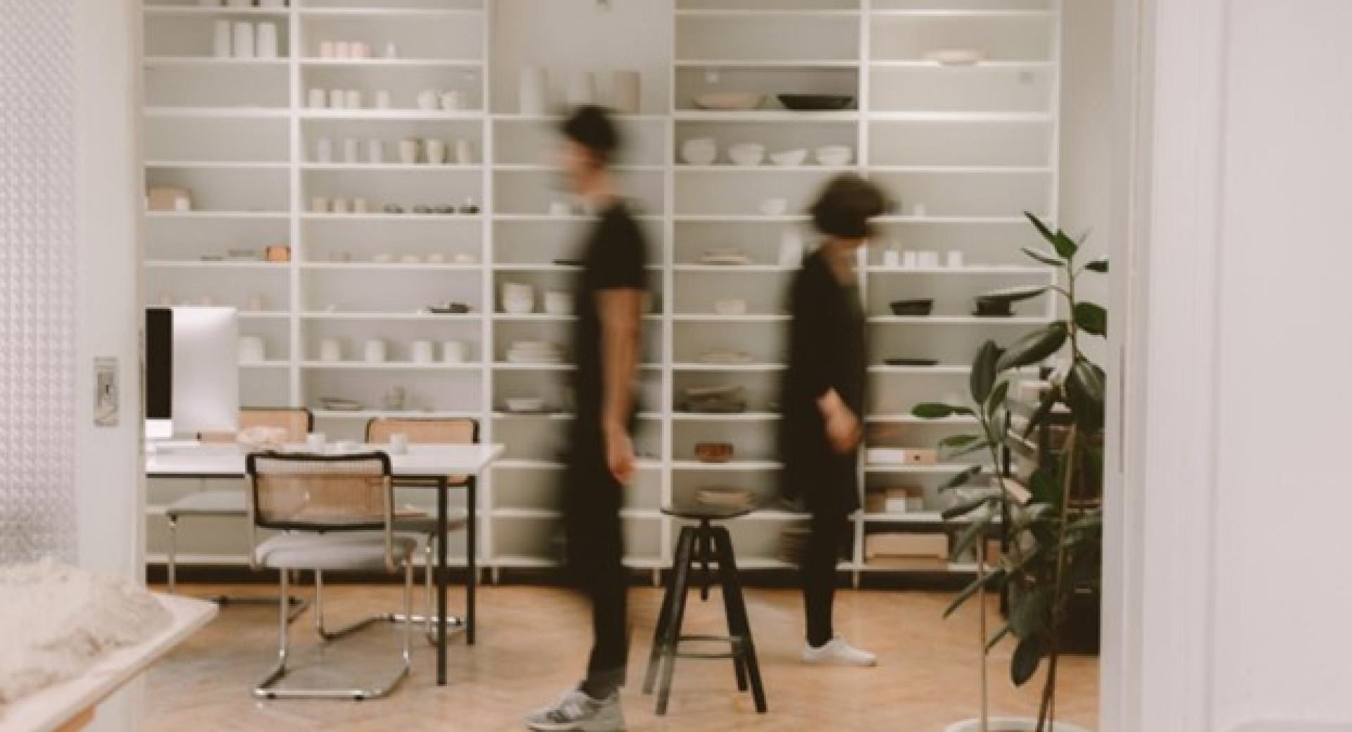 Spacious modern room with white shelves filled with various tableware and ceramics. Two blurred figures walk by.