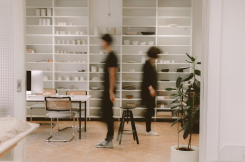 Spacious modern room with white shelves filled with various tableware and ceramics. Two blurred figures walk by.