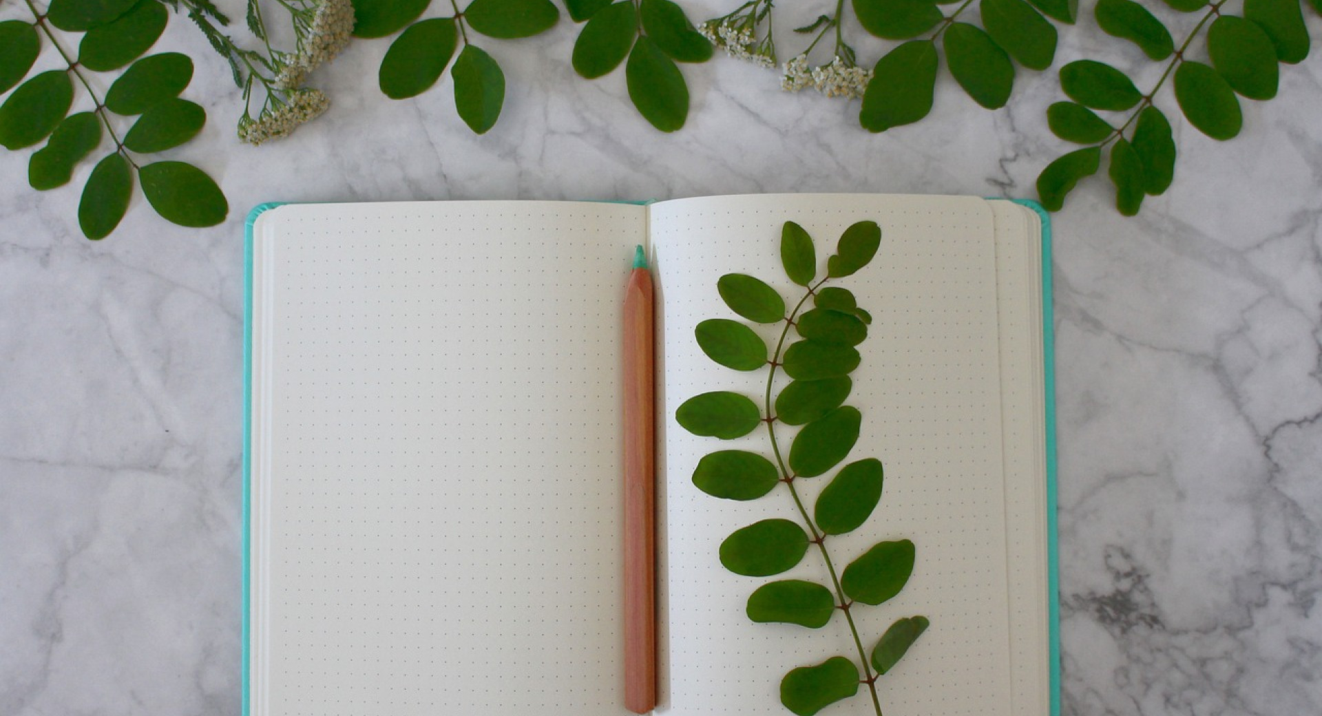 On the marble surface lies an open, dotted notebook with a turquoise cover. A light brown ink pencil is placed on the open, blank pages. Several green leaves are scattered around the notebook.