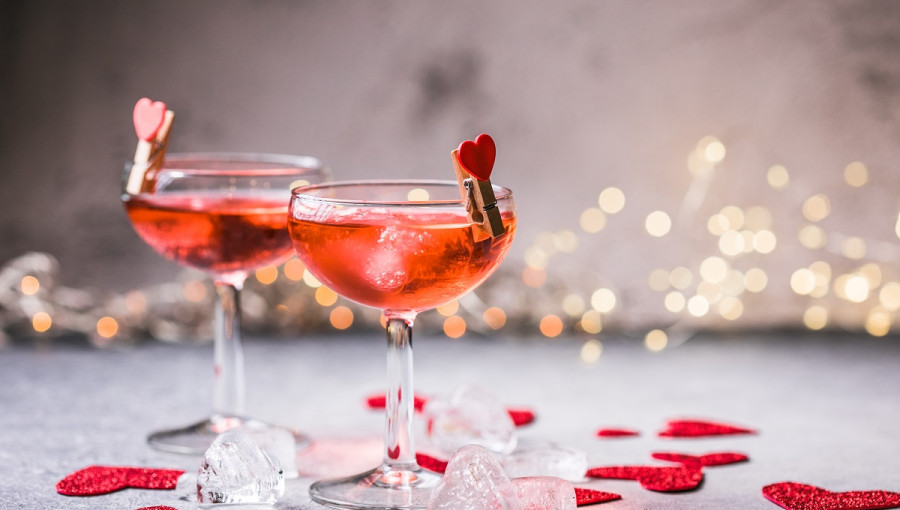 Two glasses with cocktail on a grey surface. Red hearts scattered around them.