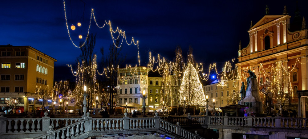 Boats on the river, with buildings and trees adorned with festive lights, glowing in vibrant colours in the evening.