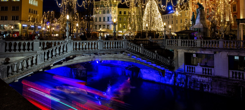 Boats on the river, with buildings and trees adorned with festive lights, glowing in vibrant colours in the evening.