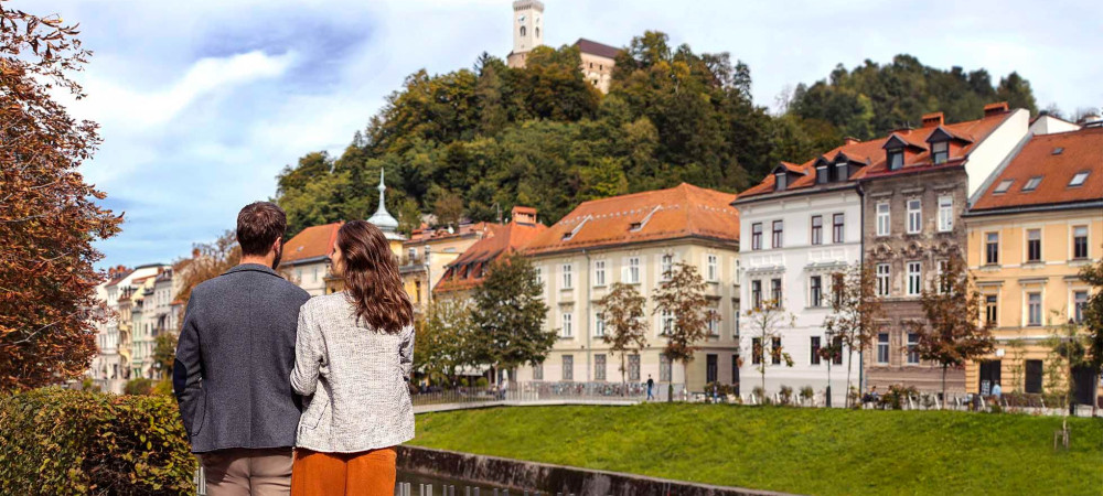 Zaljubljen par stoji na nabrežju Ljubljanice, v ozadju Ljubljanski grad.