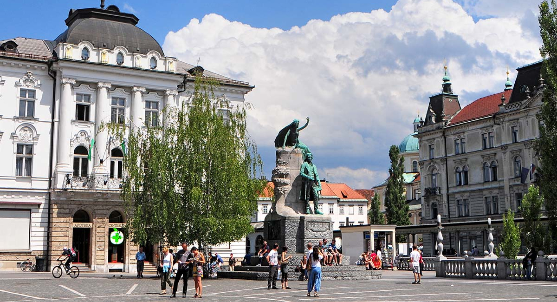 Na zivahnem trgu se nahaja velik kip figure z dvignjeno roko, ki stoji na kamnitem podstavku. V ozadju so stavbe in drevesa, ljudje pa se pod delno oblacnim nebom sprehajajo in zbirajo v blizini kipa.