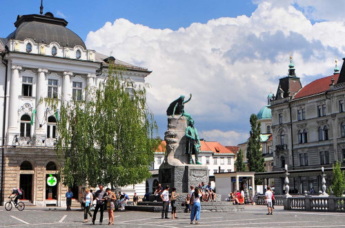 Na zivahnem trgu se nahaja velik kip figure z dvignjeno roko, ki stoji na kamnitem podstavku. V ozadju so stavbe in drevesa, ljudje pa se pod delno oblacnim nebom sprehajajo in zbirajo v blizini kipa.