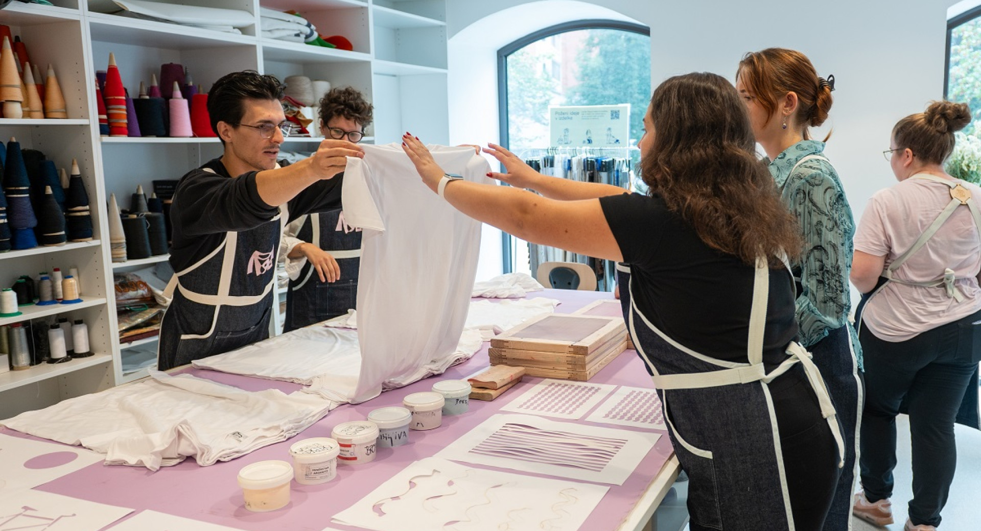 A large table with colors and patterns for printing on a t-shirt. A man is holding a white t-shirt in the air, while a group of people is watching him.