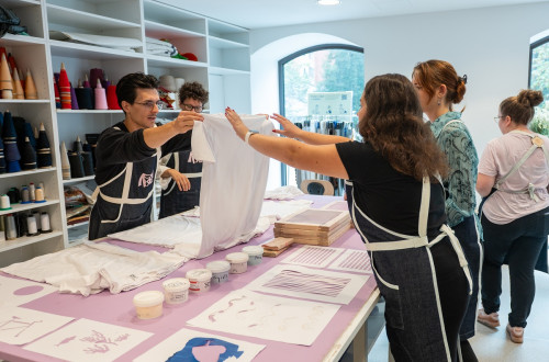 A large table with colors and patterns for printing on a t-shirt. A man is holding a white t-shirt in the air, while a group of people is watching him.